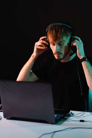 Portrait of Software Developer. Hacker. Gamer Wearing Headset Sitting at His Desk and Working. Playing on Laptop. In the Background Dark High Tech Environment with red neon lights.の写真素材