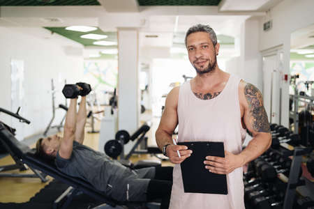 Muscular handsome trainer writing a fitness plan on clipboard for working out near his trainee in...
