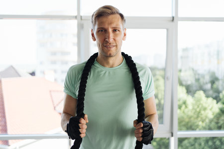 Caucasian blonde man training with rope in functional training fitness gym with big windows and a...