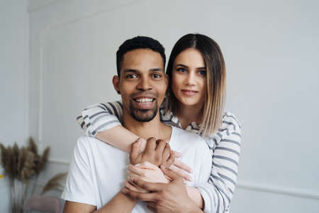 Multiracial couple in love embraces in cozy living room at home. A close-up portrait of smiling caucasian woman and african man are hugging. Love and affection conceptの写真素材