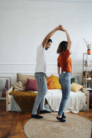 Cheerful active romantic african caucasian couple dancing in bedroom together, happy carefree young black husband and white wife enjoy weekend morning laughing bonding having fun at homeの写真素材