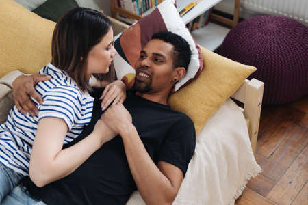 Portrait of happy young African Caucasian couple hug sitting on couch at home, smiling mixed race husband and wife spend time together, embrace relaxing on sofa. Healthy relationships conceptの写真素材