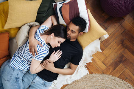 Portrait of happy young African Caucasian couple hug lying on couch at home, mixed race husband and wife spend time together, embrace sleeping on sofa. Healthy relationships conceptの写真素材