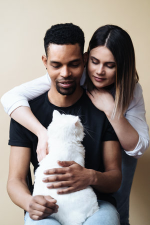 Lovely mixed-races young happy joyful couple male and female smiling in good mood resting with pet animal holding cat in hands, pet lover concept on beige backgroundの写真素材