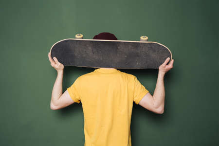 Portrait of young fashionable hipster man posing over green wall background. Caucasian male holding skateboard and smilingの写真素材