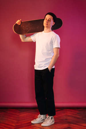 Portrait of young fashionable hipster man posing over crimson wall background splashed by neon lights. Caucasian male holding skateboard and smilingの写真素材