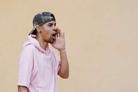 Attention. Close up portrait of cheerful young afro man in pink parka is holding hand near his open mouth isolated on pale yellow background with copy space for textの写真素材