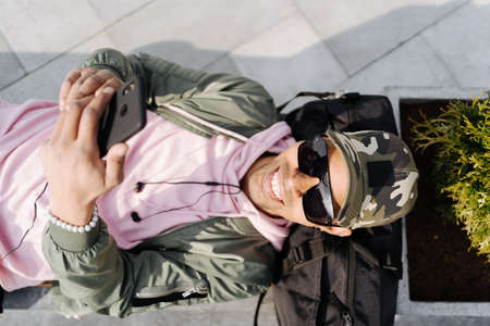 People, technology, travel and tourism - Afro man using smartphone on city street bench and texting on a sunny dayの写真素材