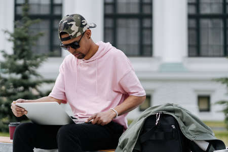 Portrait of young smiling afro man in sunglasses using laptop computer while sitting on bench outdoorsの写真素材