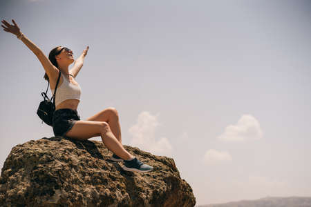 Young happy woman with backpack sitting on a rock with raised hands and looking to the blue skyの写真素材
