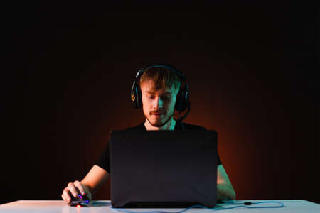 Portrait of Software Developer. Hacker. Gamer Wearing Headset Sitting at His Desk and Working. Playing on Laptop. In the Background Dark High Tech Environment with red neon lights.の写真素材