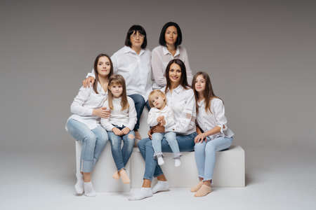 Delighted cute young girls, their moms and grandmother smiling, hugging while sitting on white cubes on gray background wearing jeans and white shirts. Lovely women groupの写真素材