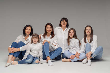 Delighted cute young girls, their moms and grandmother smiling, sitting on gray background wearing...