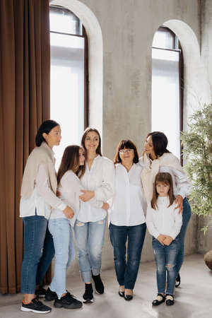 Delighted cute young girls, their moms and grandmother smiling, hugging while standing at home in the living room wearing jeans and white shirts. Lovely women groupの写真素材
