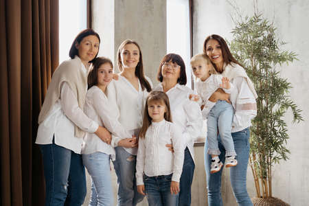 Delighted cute young girls, their moms and grandmother smiling, hugging while standing at home in...