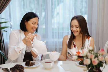 Woman pouring tea from a white teapot in white cup at home while sitting at the table and serving sisters smilingの写真素材