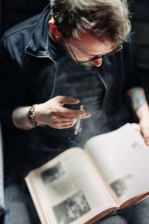 Adult student reading a book in the college library while smoking. Young male wearing glasses with...