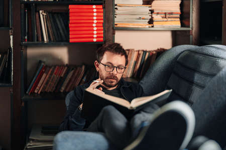 Young teacher reading a book in the college library while smoking. Young male wearing glasses with bookshelves on background sitting on couch.の写真素材
