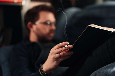 Man smoking and reading, holding a book and a cigarette in hands while lying on a couch. Focus on cigarette.の写真素材