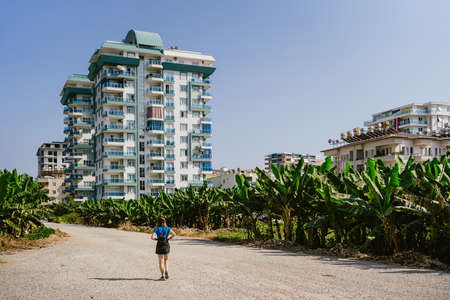 Woman walking among banana plantation on a sandy path leading to a new real estate building with sky in backgroundの写真素材