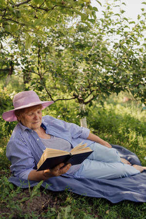 Senior woman smiling and reading a book while lying and relaxing on a blanket in the green gardenの写真素材