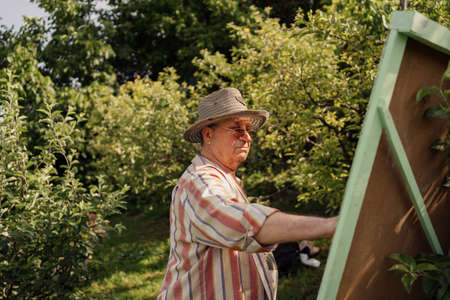 Senior man wearing a hat, painting in his garden among green trees