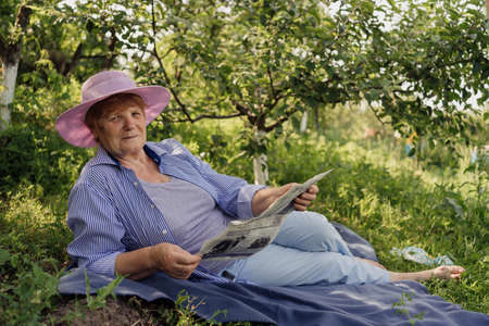 Senior woman smiling and reading a book while lying and relaxing on a blanket in the green gardenの写真素材