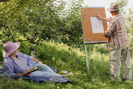 HAPPY SENIOR COUPLE spend time together in their garden or nature. A man is painting his wife while she is relaxing with a book. Growing old beautifully together. Vibrant green gardenの写真素材
