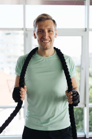 Caucasian blonde man training with rope in functional training fitness gym with big windows and a...