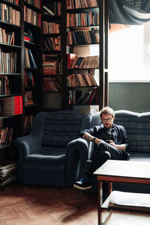 Man wearing glasses using a smartphone while sitting on a couch in the library with books in the...