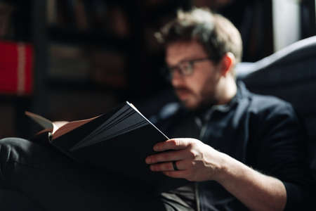 Adult student reading a book in the college library. Young positive male wearing glasses with bookshelves on background.の写真素材