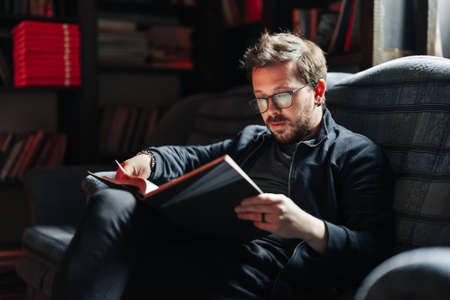 Adult student reading a book in the college library. Young positive male wearing glasses with bookshelves on background.の写真素材