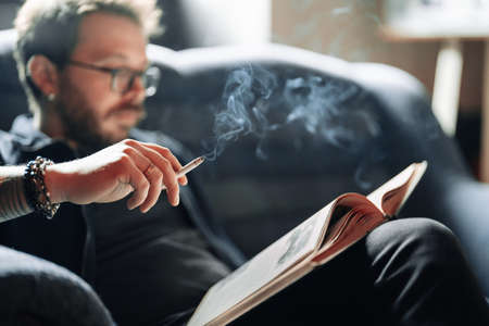 Adult student reading a book in the college library while smoking. Young male wearing glasses with bookshelves on background sitting on couch.の写真素材