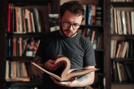 Adult student reading a book in the college library. Young positive male wearing glasses with bookshelves on background.の写真素材