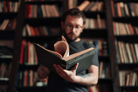 Adult student reading a book in the college library. Young positive male wearing glasses with bookshelves on background.の写真素材