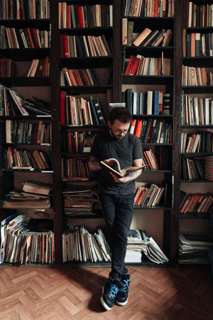 Adult student reading a book in the college library. Young standing male wearing glasses with bookshelves on background.の写真素材