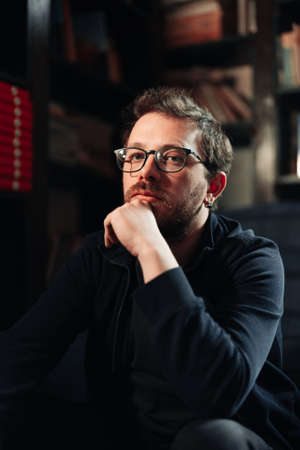 Positive male student wearing glasses in library with bookshelves on background. Proud and...