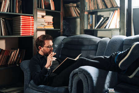 Young teacher reading a book in the college library while smoking. Young male wearing glasses with bookshelves on background sitting on couch.の写真素材