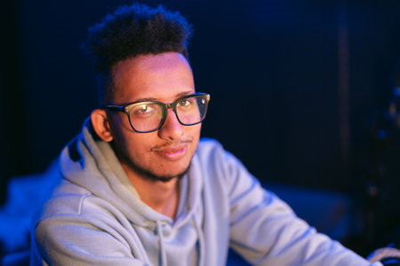 Dark skinned young man with a beaming smile, wears a gray parka, and glasses on dark background...