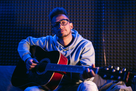 Young afro musician wearing glasses playing acoustic guitar, on dark backgroundの写真素材