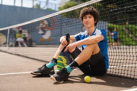 Portrait of sporty young man with curly hair posing on the padel outdoor court with a racket and a ball. Spots concept, healthy lifestyleの写真素材