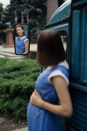 Pretty pregnant smiling woman in a retro blue dress with white collar looking in the mirror of a vintage busの写真素材