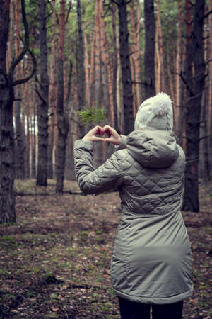 A man in light clothes with a white hat stands in the forest and shows a heart with his fingers. A man in light clothes and a white hat, in the forest, shows his love for nature.の写真素材