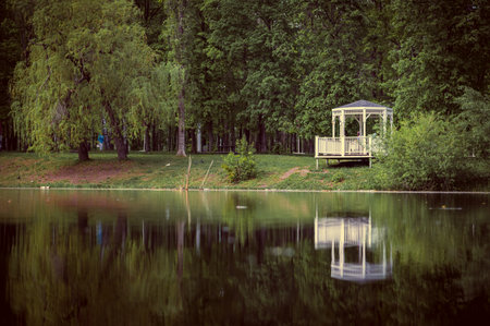 A white rotunda, with a green roof, in the park, on the shore of a small quiet pond, in summer. A quiet, small pond, with a beautiful white rotunda, is reflected in the pond.の写真素材