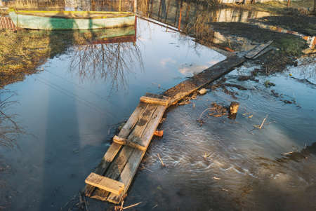 Plank boards folded in the bridge across a stream during the spring flood highwater against the background of a boat, fence of the house, in town.の写真素材