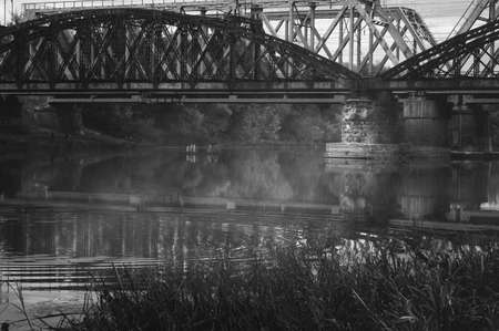 Black and white image of old railway bridges over the autumn foggy river. Gloomy atmospheric cityscapeの写真素材