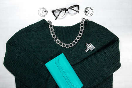 On a white background lies a dark green women's sweater, a large metal chain, transparent glasses, earrings and a turquoise large women's walletのeditorial素材