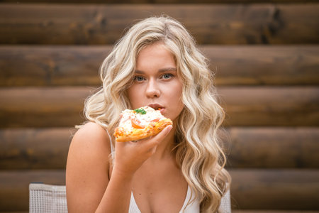 Beautiful girl with blond wavy hair in white clothes eating pizza on a wooden wall backgroundの写真素材