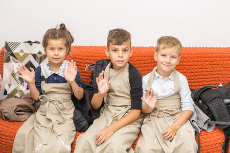 Three children are sitting in aprons on a red sofa and waving their hands at the cameraの写真素材