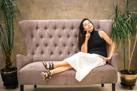Woman with dark hair in a white skirt sits on the sofa and smiles.の写真素材